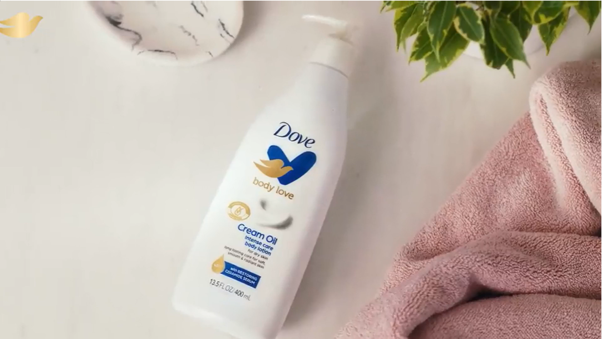 Frasco da loção Dove Body Love Cream Oil ao lado de toalhas cor-de-rosa e folhas verdes.
