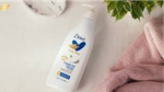 Frasco da loção Dove Body Love Cream Oil ao lado de toalhas cor-de-rosa e folhas verdes.