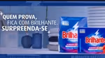 Duas embalagens do produto de limpeza Brilhante em destaque contra um fundo azul, com roupas dobradas e utensílios de limpeza.